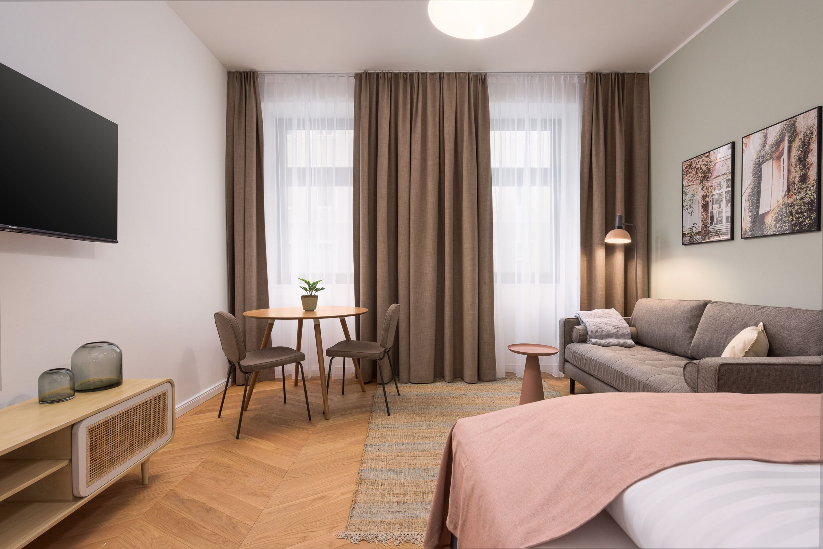 Ein modernes Hotelzimmer mit Flachbildfernseher, einer Couch, einem Bett, einer Essecke mit Stühlen und einem Tisch sowie großen Fenstern mit Vorhängen.