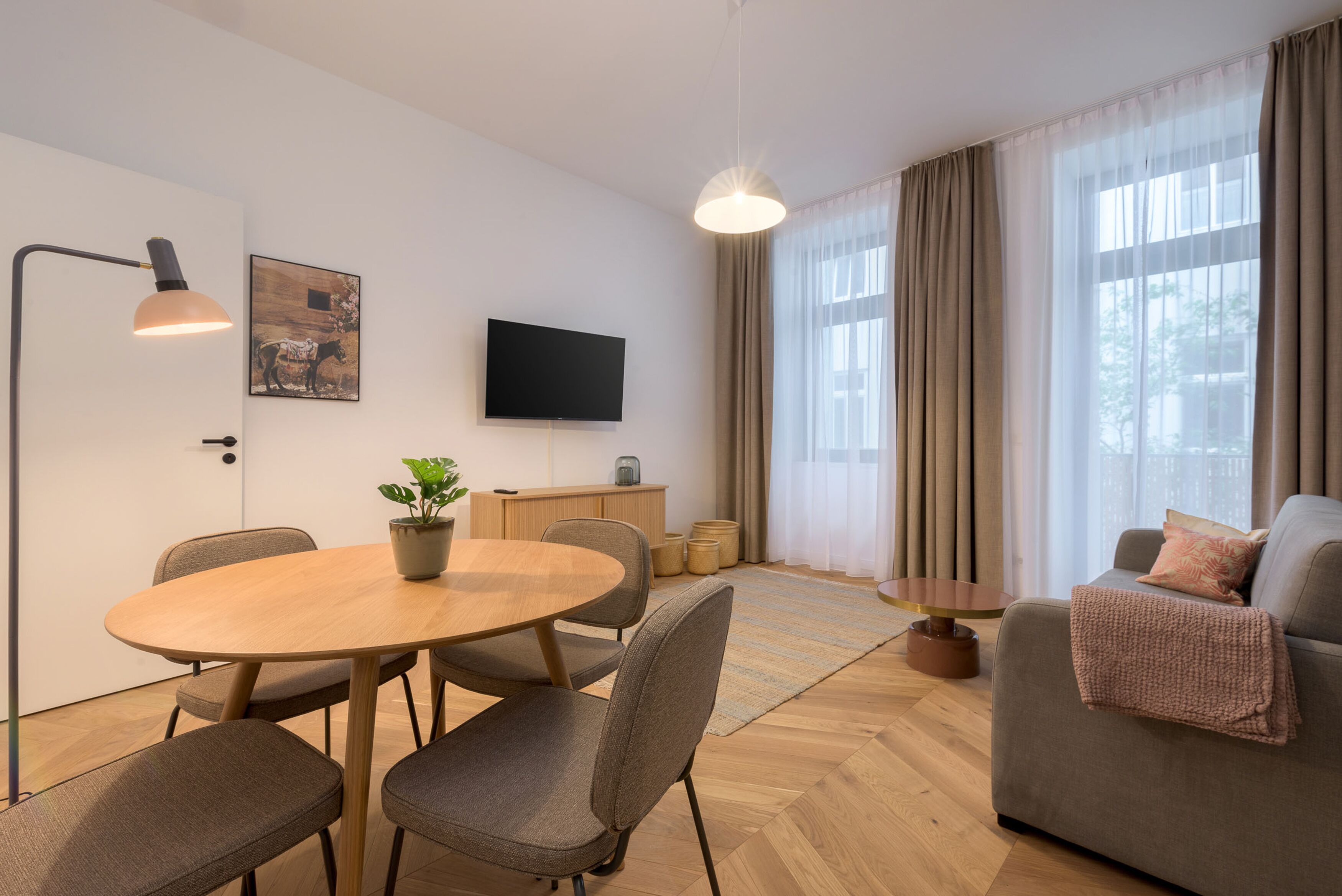 Un soggiorno moderno con un tavolo rotondo in legno, sedie grigie, una divano, una TV a parete e un'ampia finestra con tende.