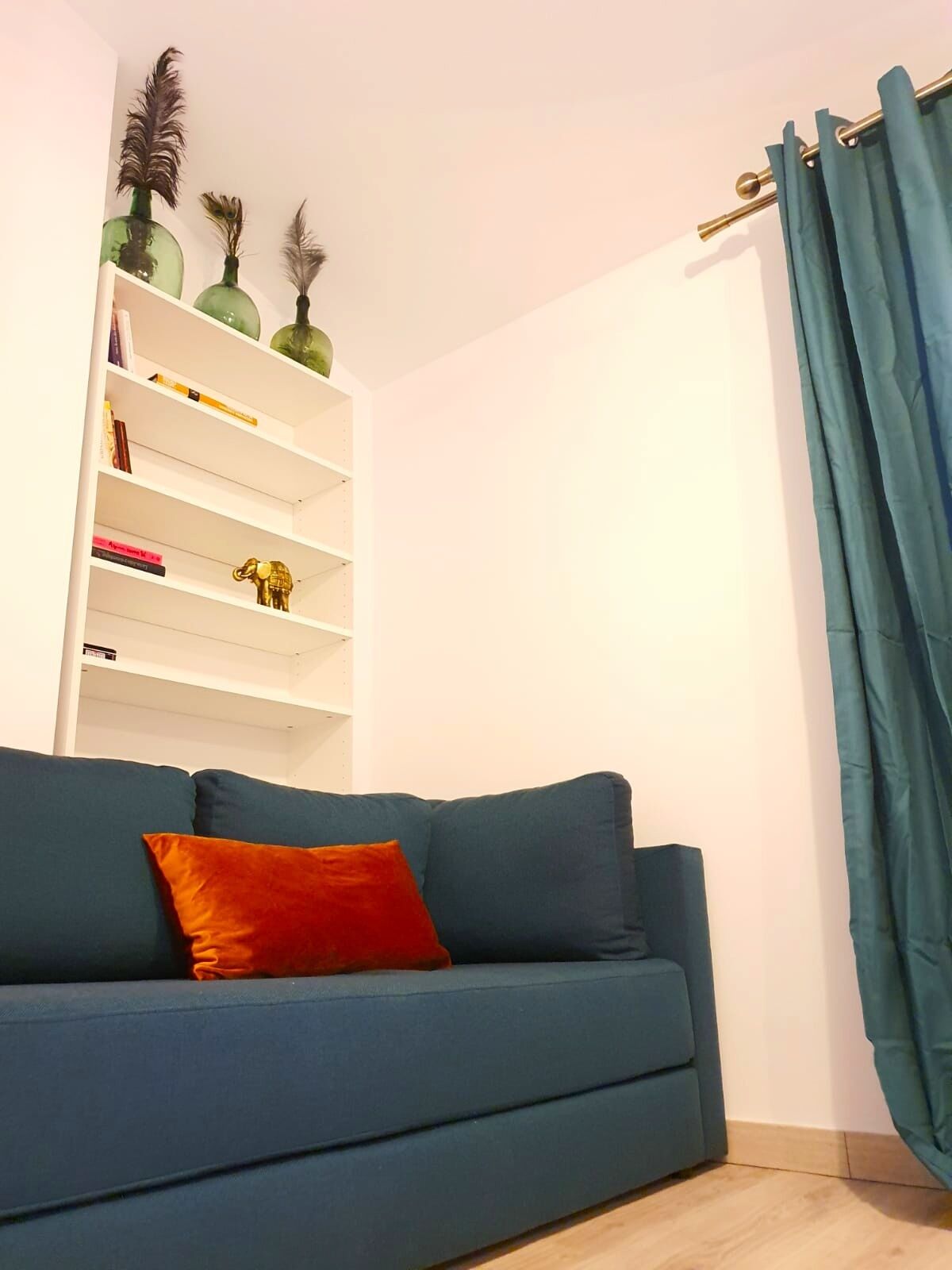Una habitación con un sofá azul, una estantería blanca con adornos y una cortina color turquesa.