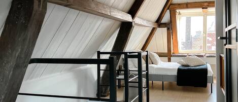 A modern loft bedroom with a sloped ceiling, exposed wooden beams, a glass bed frame, and a view of the city through large windows.