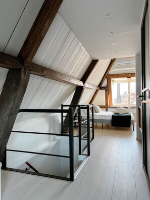 Un dormitori tipus loft modern amb un sostre inclinat, bigues de fusta al descobert, un llit amb estructura de vidre i vista a la ciutat a través de grans finestres.
