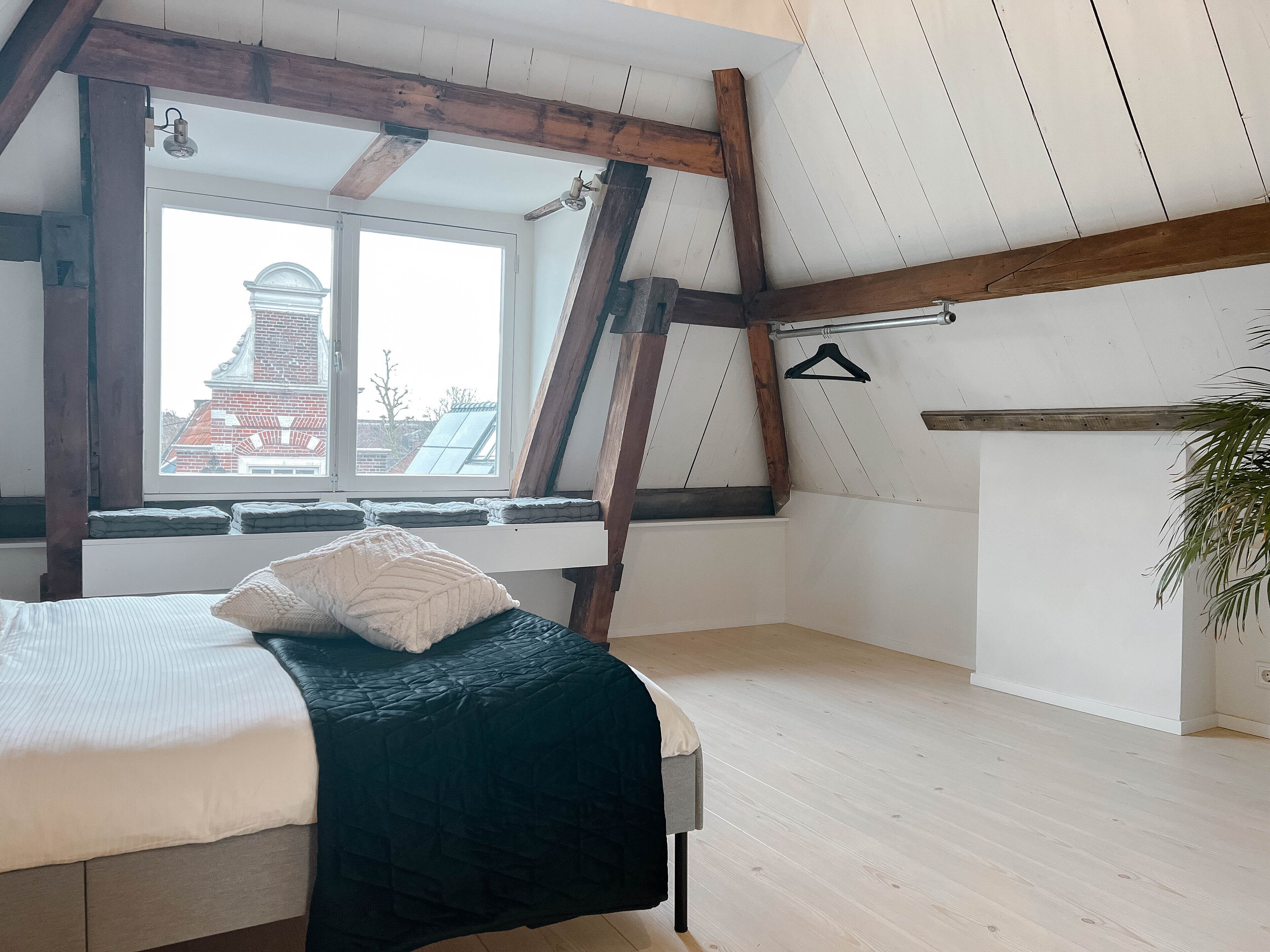 A bedroom with a bed, a window with a view of buildings, wooden beams, and a plant.