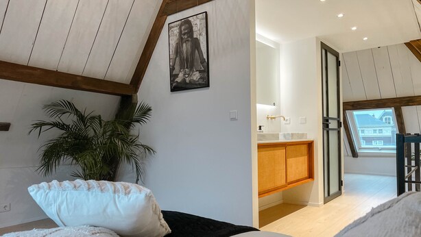 Un dormitorio con cama, almohadas, una planta y vistas a una cocina y un balcón.