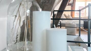 A glass vase with dried flowers on a white table, two white candles, and a view of a balcony with a sofa and a basket.