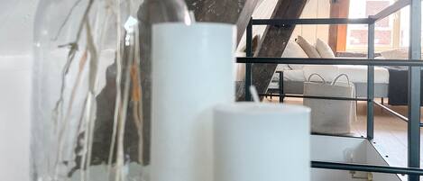 Un florero de vidrio con flores secas sobre una mesa blanca, dos velas blancas y vistas a un balcón con un sofá y una cesta.