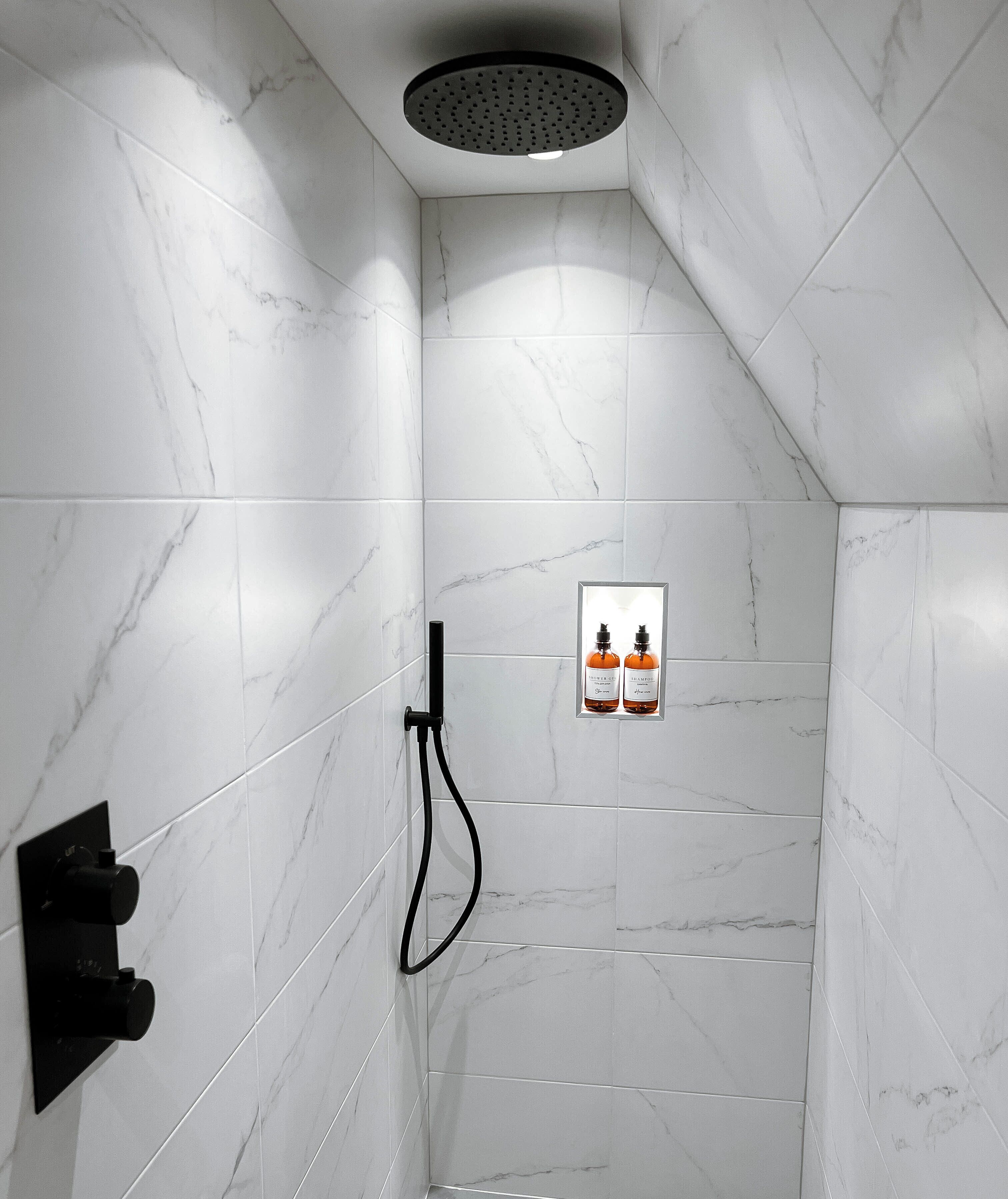A modern shower area with white marble tiles, a handheld showerhead, and two bottles of shampoo and conditioner on the wall.