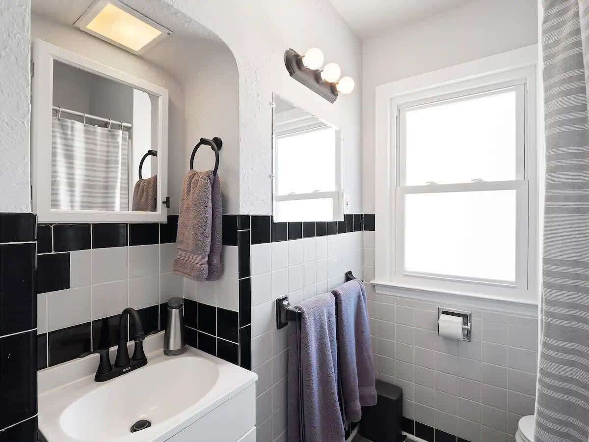 A bathroom with a white sink, black and white tiled walls, a mirror with lights, and a window.