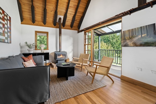 Un salon chaleureux avec une cheminée, un canapé, un fauteuil et une grande fenêtre donnant sur les arbres.
