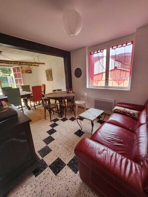 A living room with a red leather sofa, a dining table with chairs, a patterned rug, and a window with curtains.