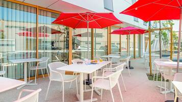 An outdoor cafe with tables and chairs, red umbrellas, and a modern building in the background.