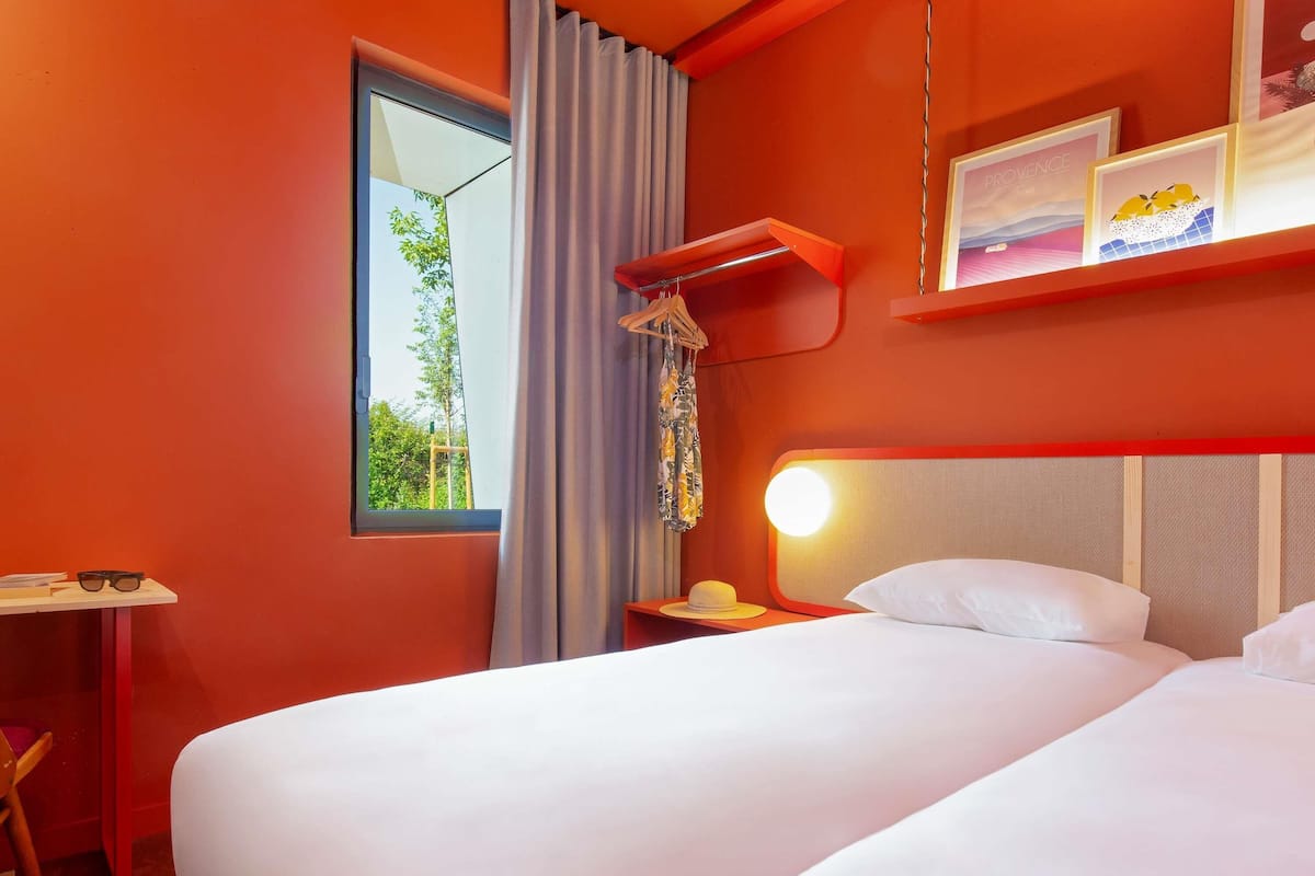 A hotel room with a large bed, a desk, a chair, a window with curtains, and a wall-mounted shelf with framed pictures.