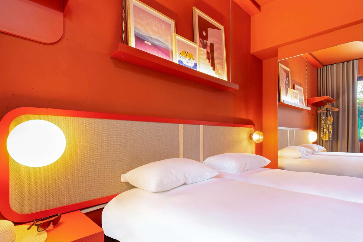 A hotel room with two beds, a red wall, and a framed picture above the beds.