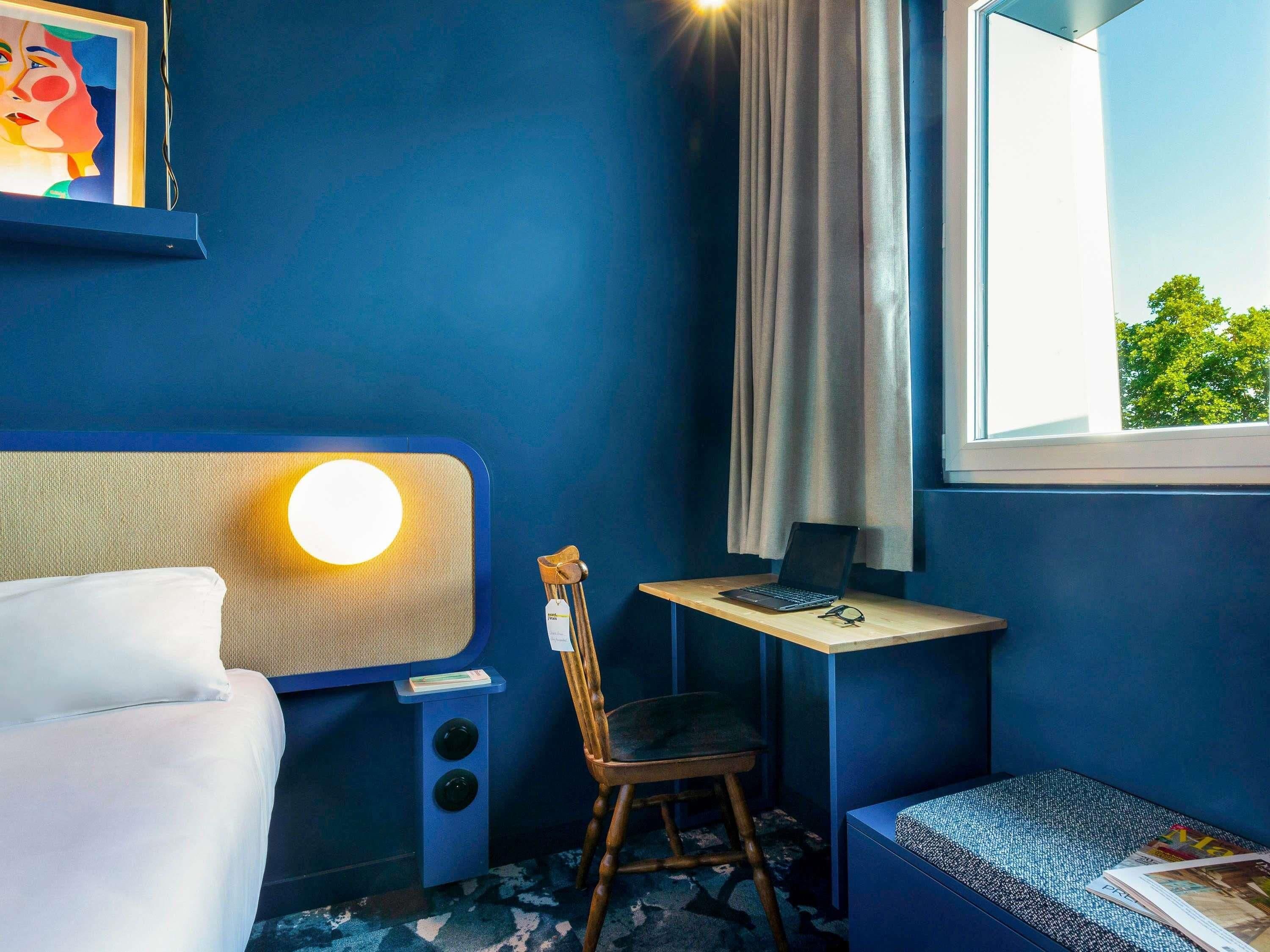 A room with a bed, a desk with a laptop, a chair, a window with curtains, and a wall-mounted lamp.