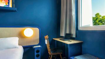 A room with a bed, a desk with a laptop, a chair, a window with curtains, and a wall-mounted lamp.