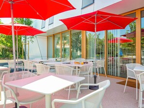 Un espace extérieur aménagé avec des tables et des chaises, des parasols rouges et de grandes fenêtres.