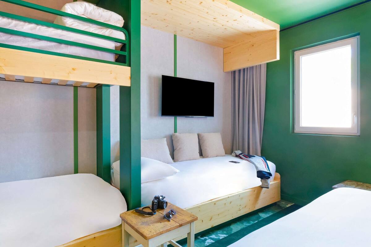A hotel room with bunk beds, a flat-screen TV, a small table, and a window with curtains.