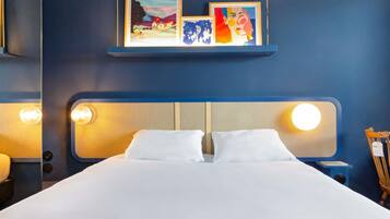Habitación de hotel con cama, dos lámparas de noche, cabecero y estante con obras de arte enmarcadas.