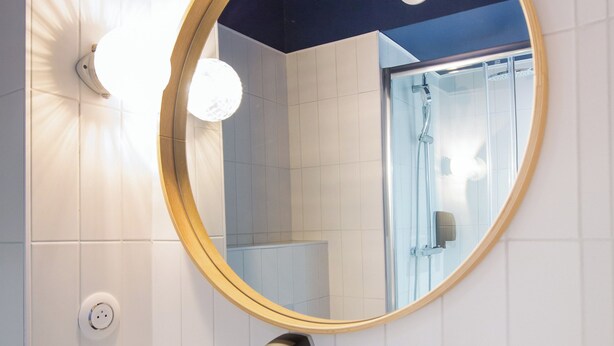 A bathroom with a round mirror, a white sink, a towel dispenser, and a shower visible in the reflection. - ibis Styles Salon de Provence (Salon-de-Provence)