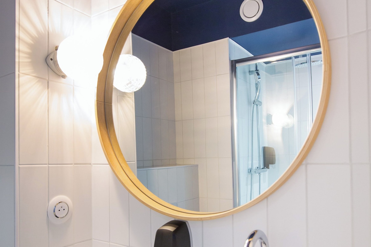 Un baño con un espejo redondo, un lavamanos blanco, un dispensador de toallas y una ducha visible en el reflejo.