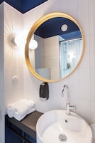 Un baño con un espejo redondo, un lavamanos blanco, un dispensador de toallas y una ducha visible en el reflejo.