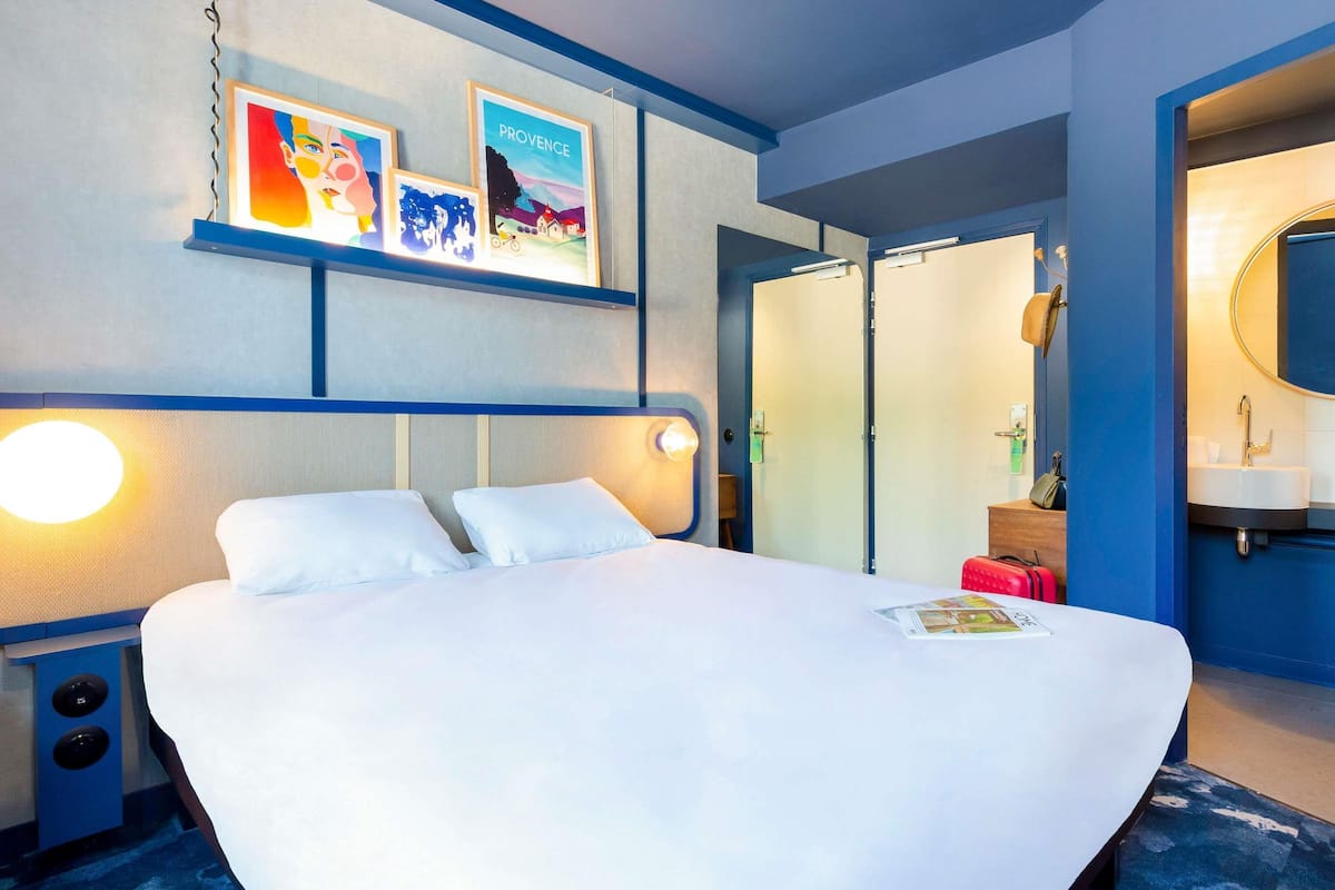 Habitación de hotel con cama, mesita de noche, televisor montado en la pared, obras de arte enmarcadas, armario y baño con espejo y lavamanos.
