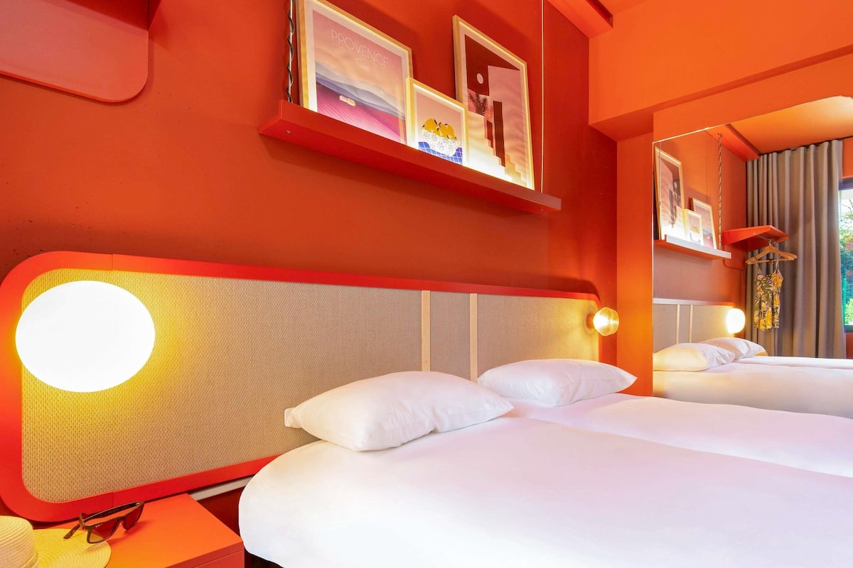 Habitación de hotel con dos camas, una pared roja y un cuadro enmarcado sobre las camas.