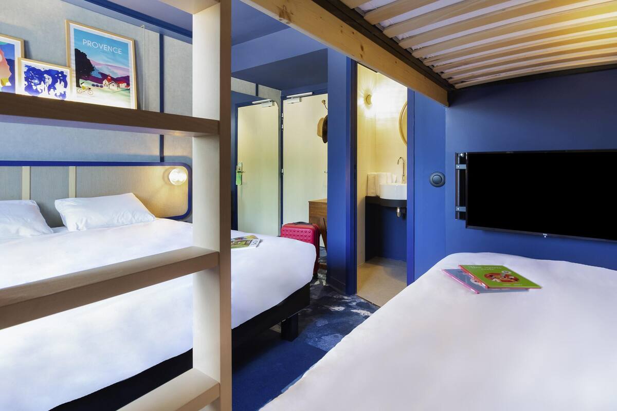 A hotel room with bunk beds, a flat-screen TV, a bathroom with a sink and mirror, and a bookshelf with travel-themed books.