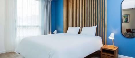 A hotel room with a large bed, a wooden headboard, a bedside table with a lamp, and a large mirror on the wall.
