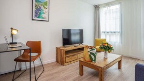 Ein modernes Wohnzimmer mit Flachbildfernseher, Schreibtisch mit Laptop, Couchtisch aus Holz, gelber Sessel und einem gerahmten Bild an der Wand.