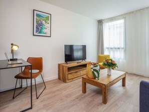 Ein modernes Wohnzimmer mit Flachbildfernseher, Schreibtisch mit Laptop, Couchtisch aus Holz, gelber Sessel und einem gerahmten Bild an der Wand.