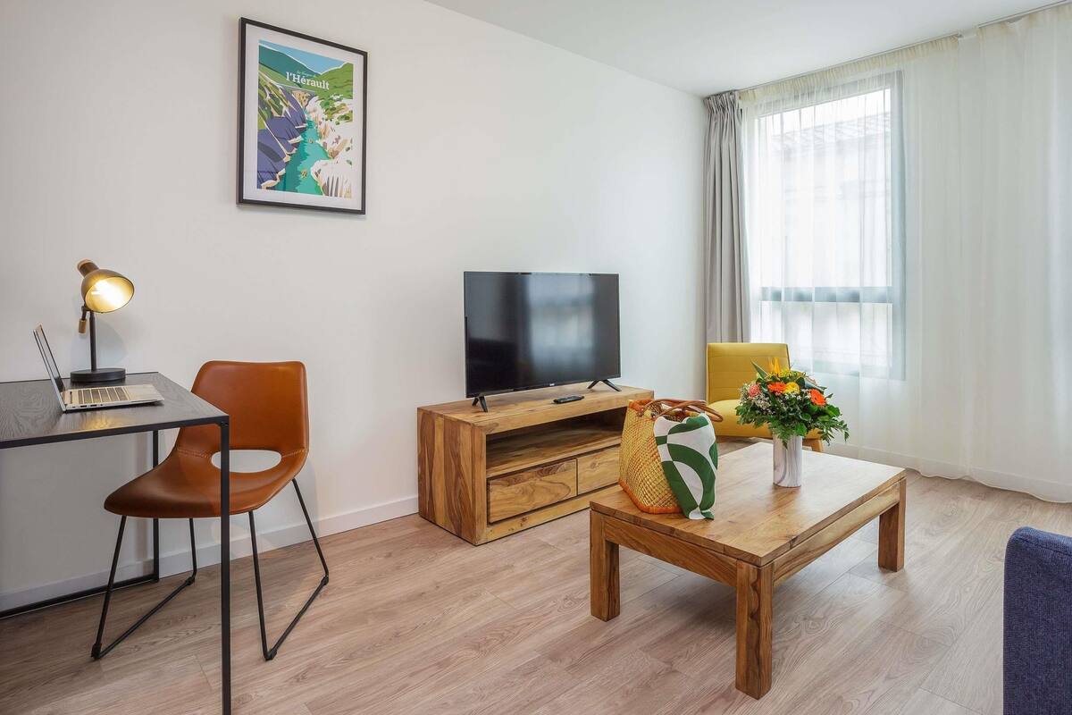Een moderne woonkamer met een houten salontafel, een televisie op een houten meubel, een laptop op een bureau, een stoel en een lijst met een foto aan de muur.