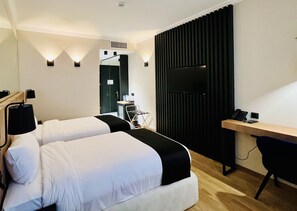 Et hotelværelse med to senge, et skrivebord, en stol, et fjernsyn og et lille tekøkken.