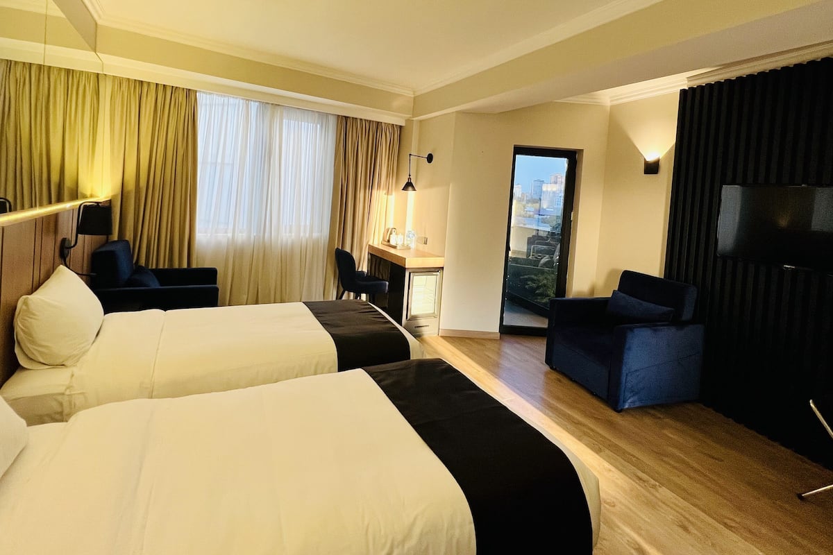 A hotel room with two beds, a desk, a chair, a TV, and a view of the city.
