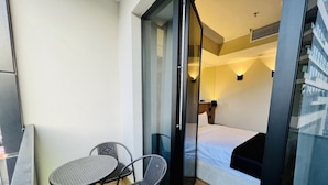 A small balcony with a table and two chairs, a bed with a wooden headboard, and a glass door leading to a room with a nightstand and lamp. - Genesis (Tbilisi)