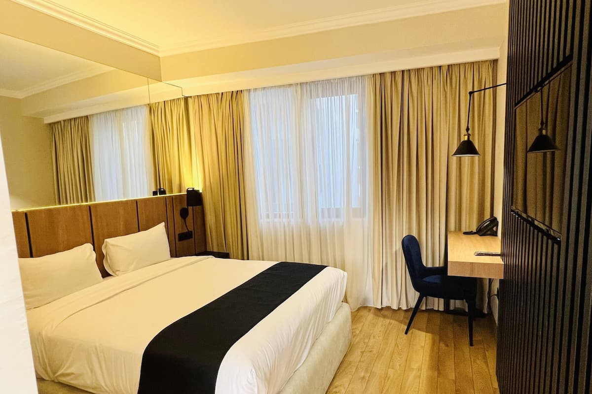 A hotel room with a large bed, a desk with a chair, and a window with curtains.
