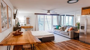 A modern living room with a wooden dining table, a sofa, and a balcony with a view. - Beachfront Condo with Amazing View (Hauula)