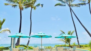 A beachside patio with three turquoise umbrellas, white chairs, and tall palm trees. - Beachfront Condo with Amazing View (Hauula)