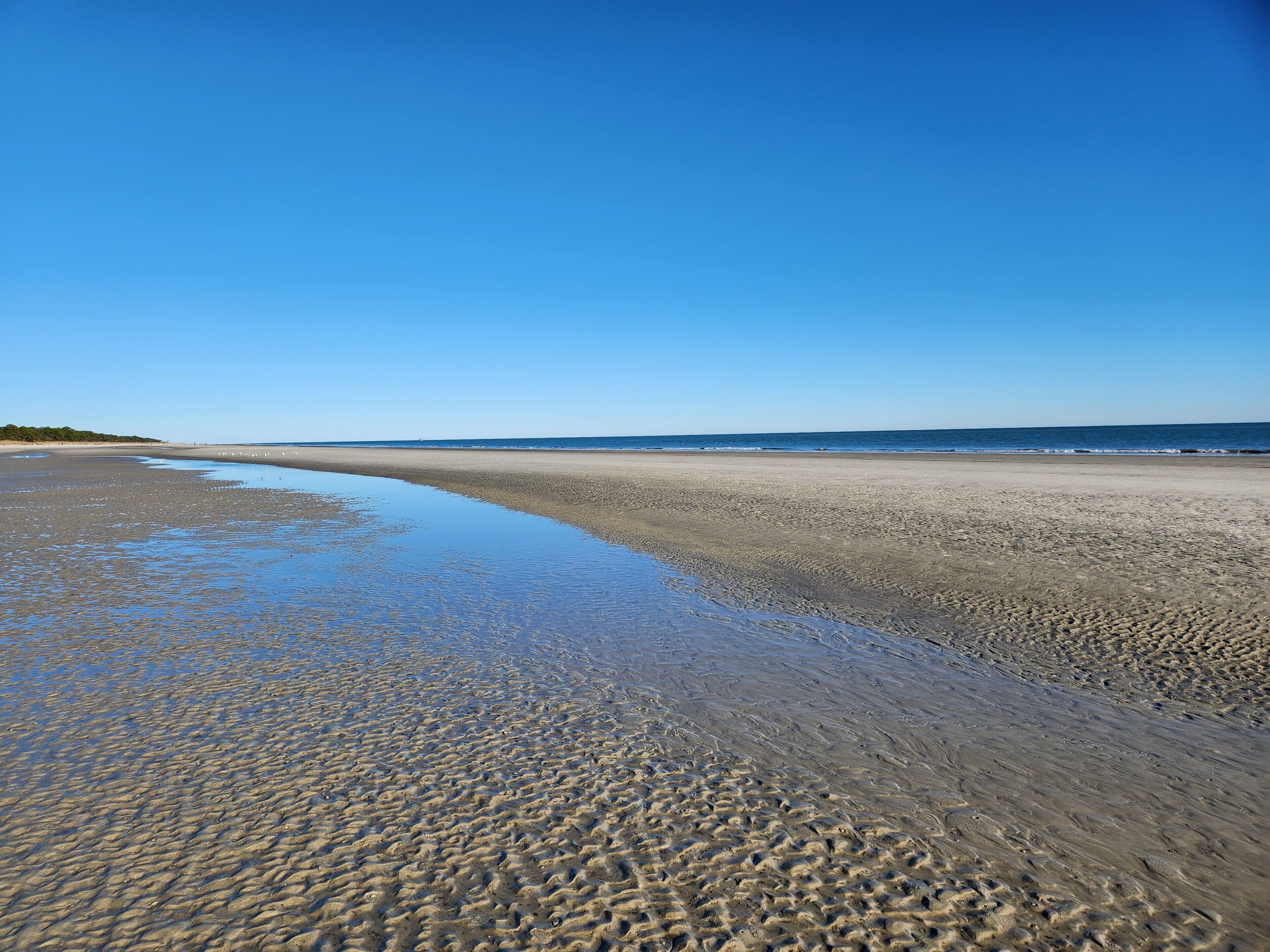 A wide, sandy beach with gentle waves lapping at the shore under a clear blue sky.