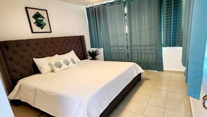 Uma cama bem arrumada com cabeceira marrom, um quadro na parede e um vaso de planta ao lado.