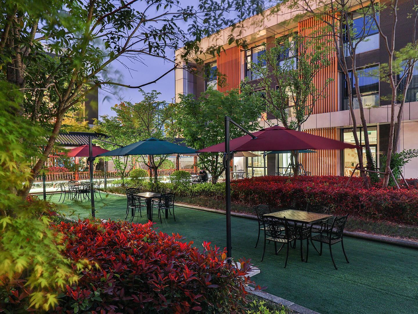 An outdoor seating area with tables and chairs under umbrellas, surrounded by lush greenery and a building with large windows in the background.