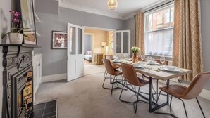 A dining area with a table set for four, a fireplace, and a view of a room with a bed and a window. - Stylish 3 Bdr Home Central Windsor By Tempstay (Berkshire)
