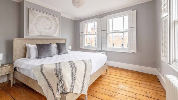 A bedroom with a wooden floor, a bed with white bedding, a white headboard, a wall-mounted artwork, and a window with shutters. - Stylish 3 Bdr Home Central Windsor By Tempstay (Berkshire)
