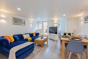 A modern living room with a blue sofa, wooden coffee table, and a dining area with a TV and plants. - Central Windsor Modern Garden Flat - By Tempstay (Berkshire)