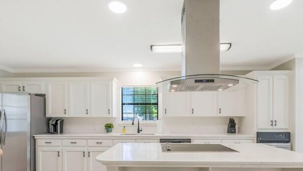 A modern kitchen with white cabinets, a central island with stools, a built-in oven, and a window with blinds.