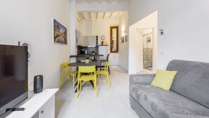 Un espacio habitable compacto con televisor, una mesa de comedor con sillas, un sofá gris con un cojín amarillo y una zona de cocina al fondo.