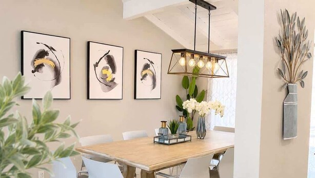 Un comedor moderno con una mesa de madera, sillas blancas, obras de arte enmarcadas, una lámpara colgante y un jarrón con plantas secas.