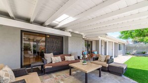 Un patio techado con mobiliario exterior, una puerta de cristal que da a un espacio interior y un césped bien cuidado.