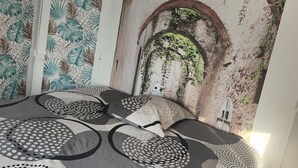 Un dormitorio con una cama con colcha de estampado geométrico, un mural de un arco con follaje y una puerta blanca con motivo de hojas.