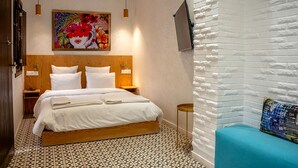 Une chambre d’hôtel avec un lit en bois, du linge de lit blanc, une œuvre d’art florale aux couleurs vives au-dessus du lit, un téléviseur fixé au mur et un mur en briques blanches servant d’élément décoratif.