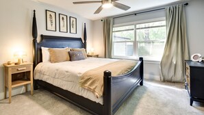 Un dormitorio con cama, mesitas de noche, ventilador de techo, ventana con cortinas y cuadros en la pared.